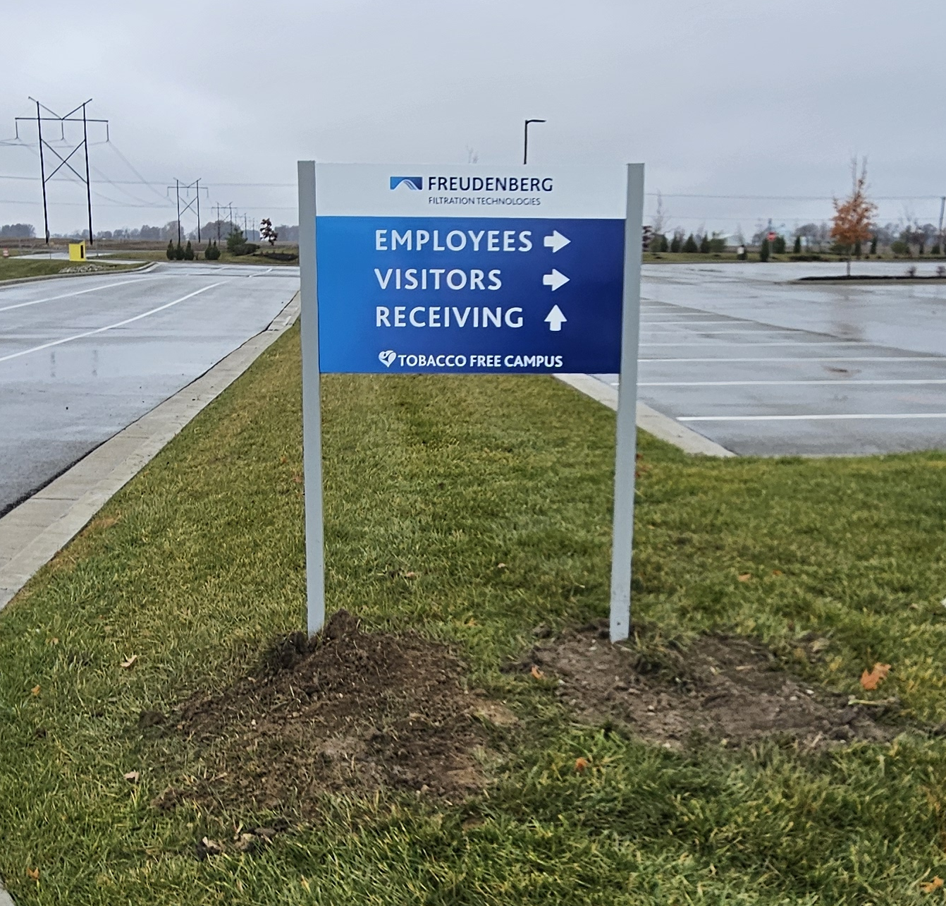Sign in a parking lot indicating directions for employees, visitors, and receiving at Freudenberg Filtration Technologies, with a message that the campus is tobacco free, on a cloudy, rainy day.