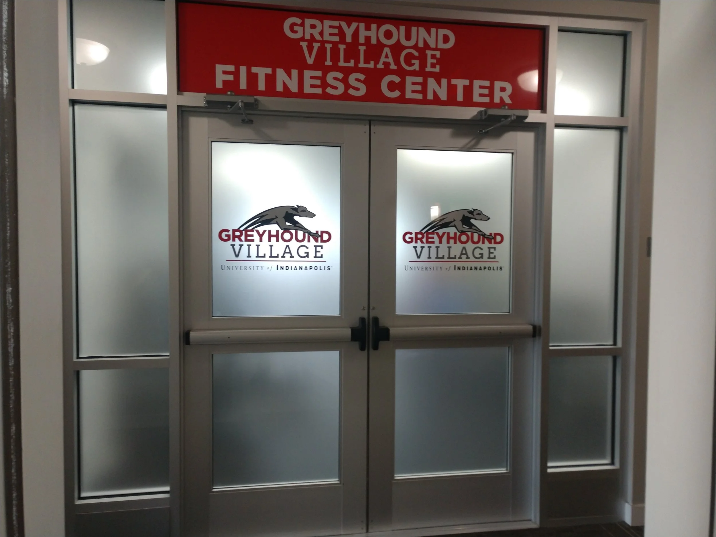Glass double doors with logos of Greyhound Village and University of Indianapolis, leading into the Greyhound Village Fitness Center.
