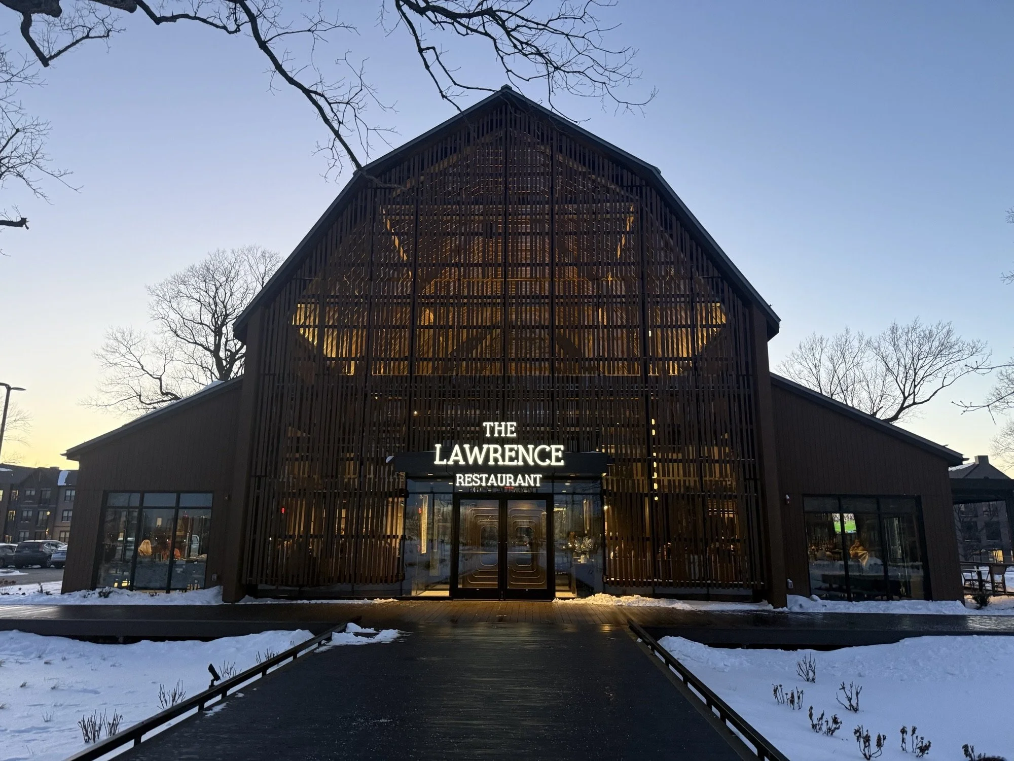 Exterior of a barn-style building with wooden slats and large glass windows, sign reading 'The Lawrence Restaurant' above entrance, snow on ground, leafless trees around, early evening lighting.