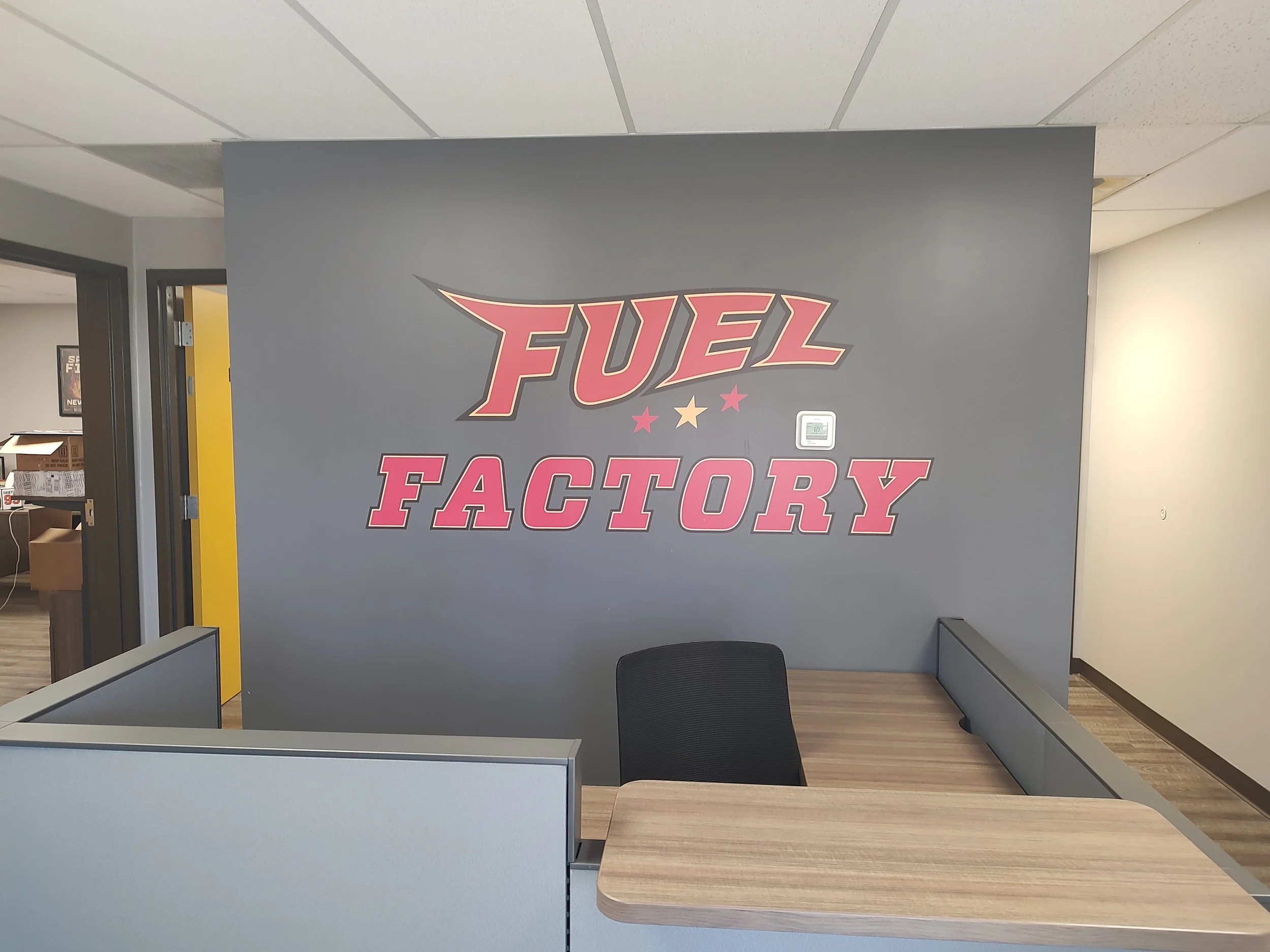 Interior of Fuel Factory office with gray wall, large pink and gold logo, black chair, and a desk.