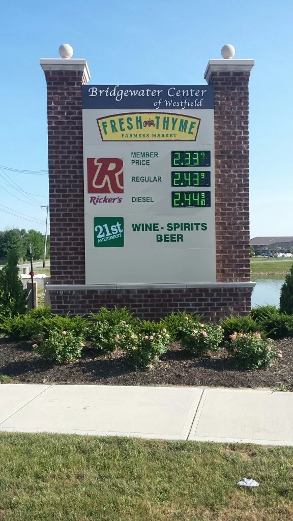 Sign for Bridgewater Center of Westfield advertising fresh thyme farmers market, with Ricker's gas station prices and a list of items including wine, spirits, and beer, in front of a sidewalk and landscaped bushes.