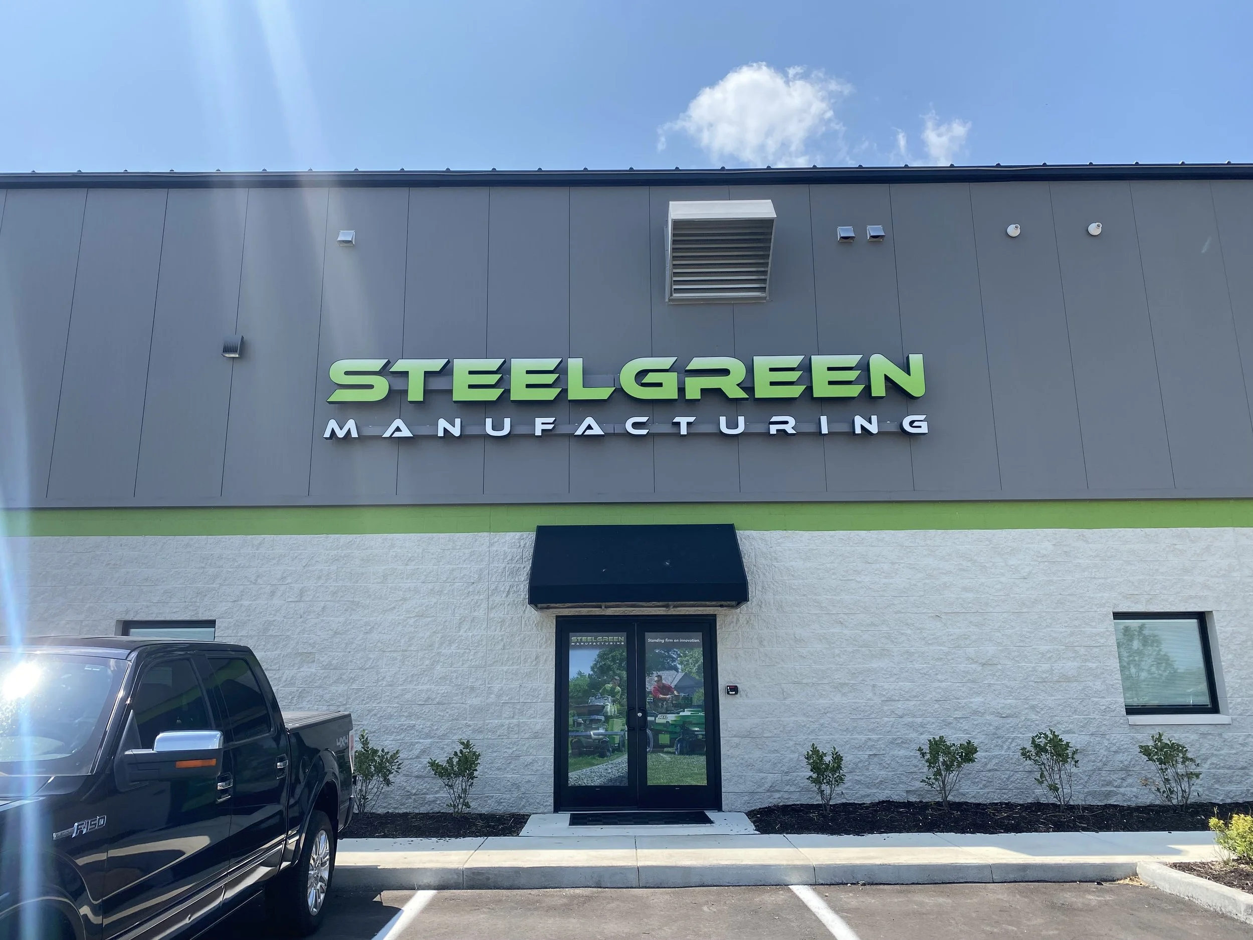 The exterior of a building with a sign reading 'Steel Green Manufacturing' in green and white letters, with a parking lot and a black truck parked in front, under a blue sky with some clouds.