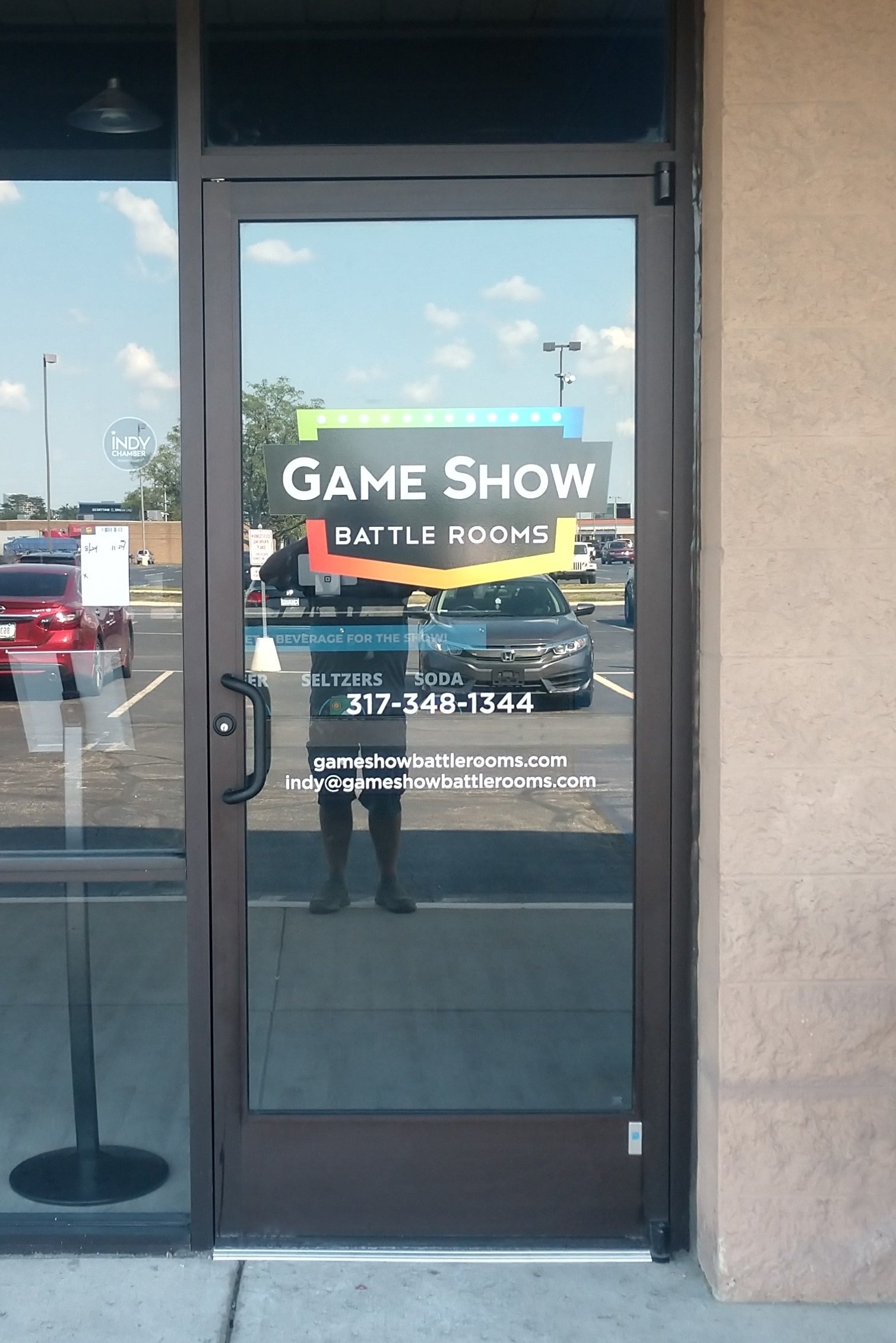 Glass door with a sign for Game Show Battle Rooms, including contact information and website, reflecting a parking lot and sky outside.
