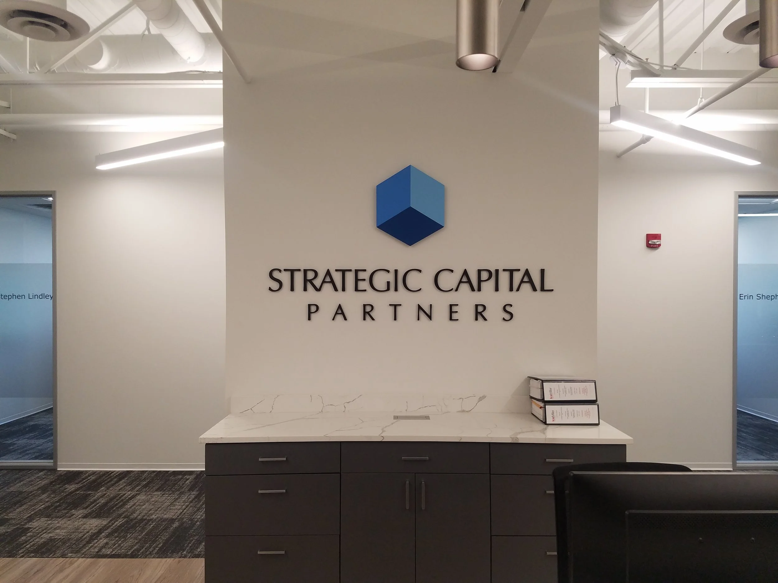 Office reception area with a white marble counter, dark drawers, and a wall sign that reads 'Strategic Capital Partners' with a blue hexagon logo above it.