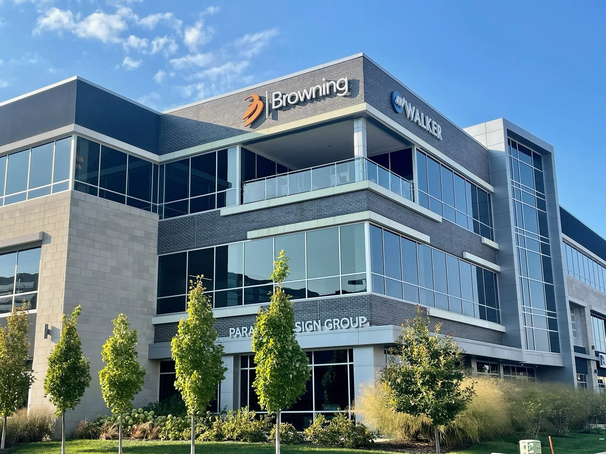 Modern multi-story office building with company signs for Browning, Walker, and Parasign Group, surrounded by landscaped greenery and trees under a blue sky with some clouds.