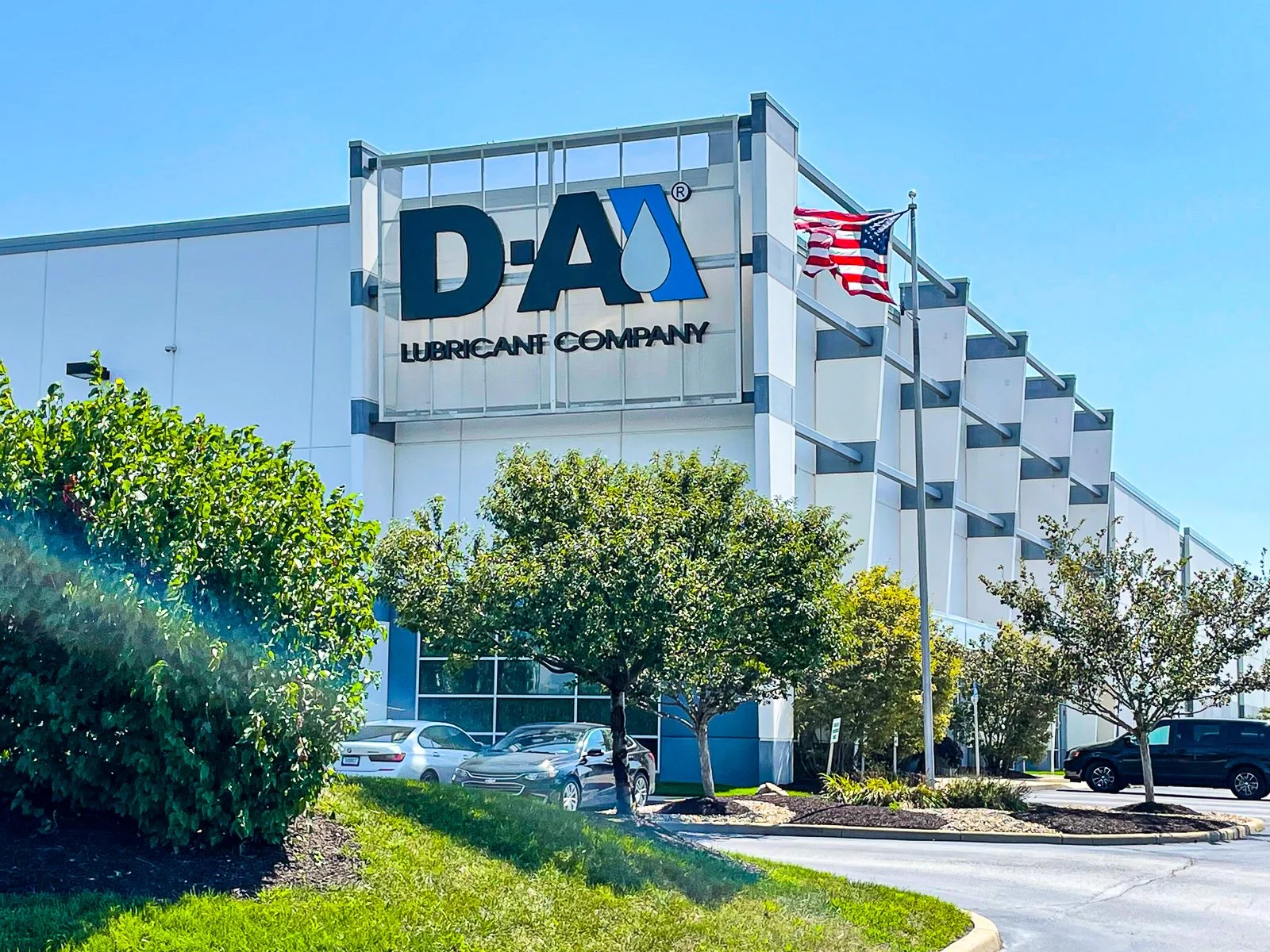 Exterior of a D-A Lubricant Company building with American flags, parking lot, trees, and a clear blue sky.