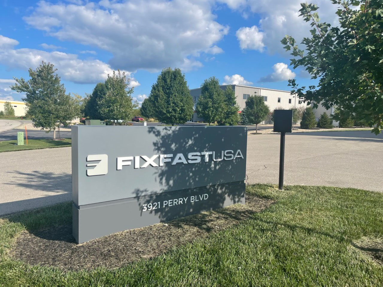 Business sign for FIXFAST USA at 3921 Perry Blvd, with trees, a parking lot, and a blue sky with clouds in the background.