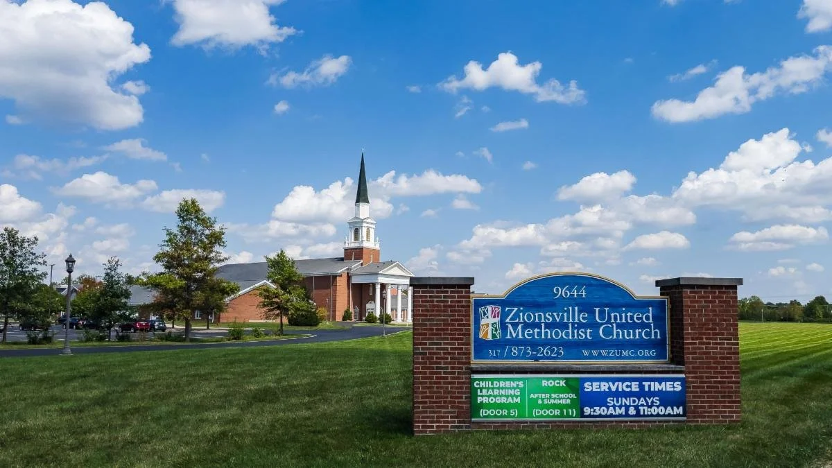 A church building with a tall steeple and a sign in front of it displaying the name Zionsville United Methodist Church, along with service times and other information, under a partly cloudy sky.
