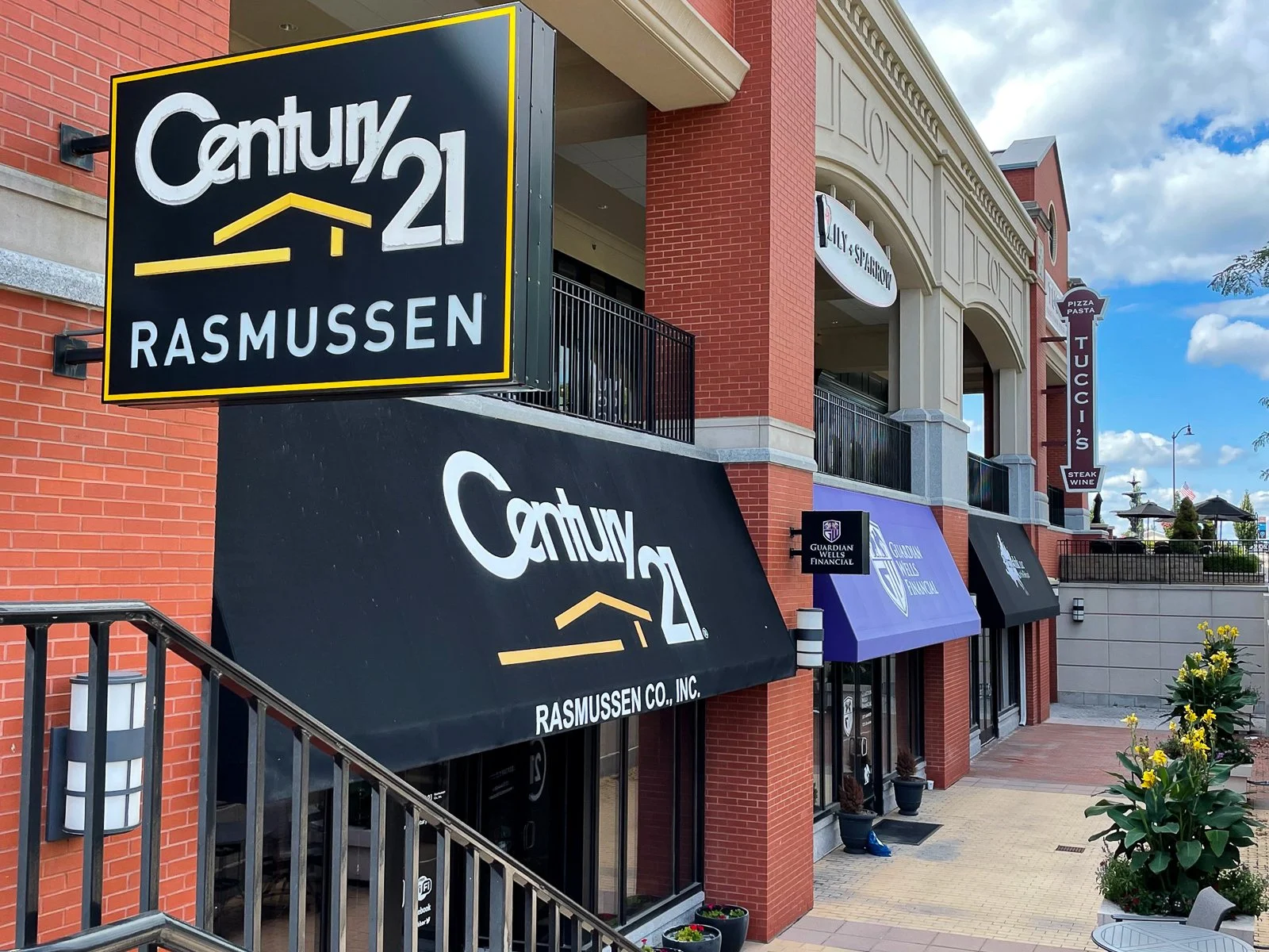Exterior view of a shopping center with businesses, including Century 21 Rasmussen real estate office with black and yellow signage, and other shops with awnings, brick walls, potted plants, and a clear sky with clouds in the background.