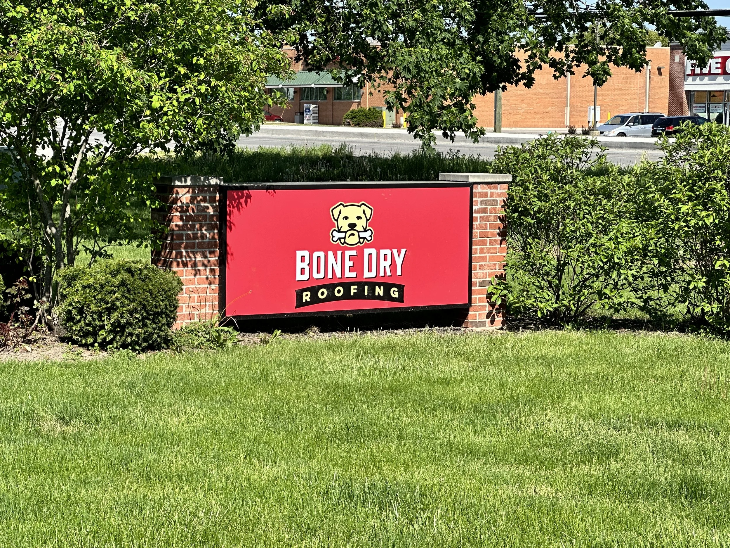 A red brick sign with green shrubbery on each side, displaying the logo and name of Bone Dry Roofing.