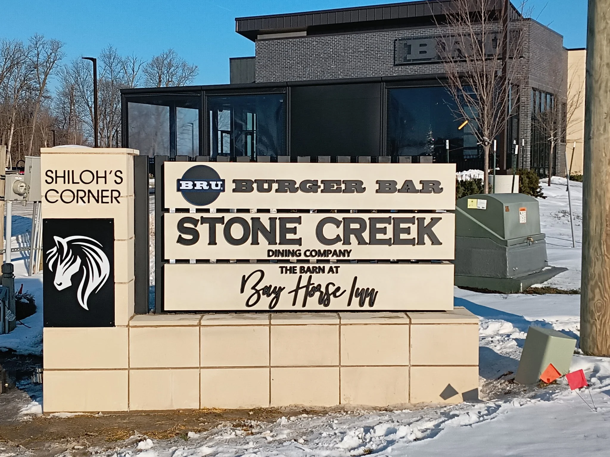 Signboard advertising BRU Burger Bar, Stone Creek Dining Company, and The Barn at Bay Horse Inn, located at Shiloh's Corner in a snowy outdoor setting with leafless trees.