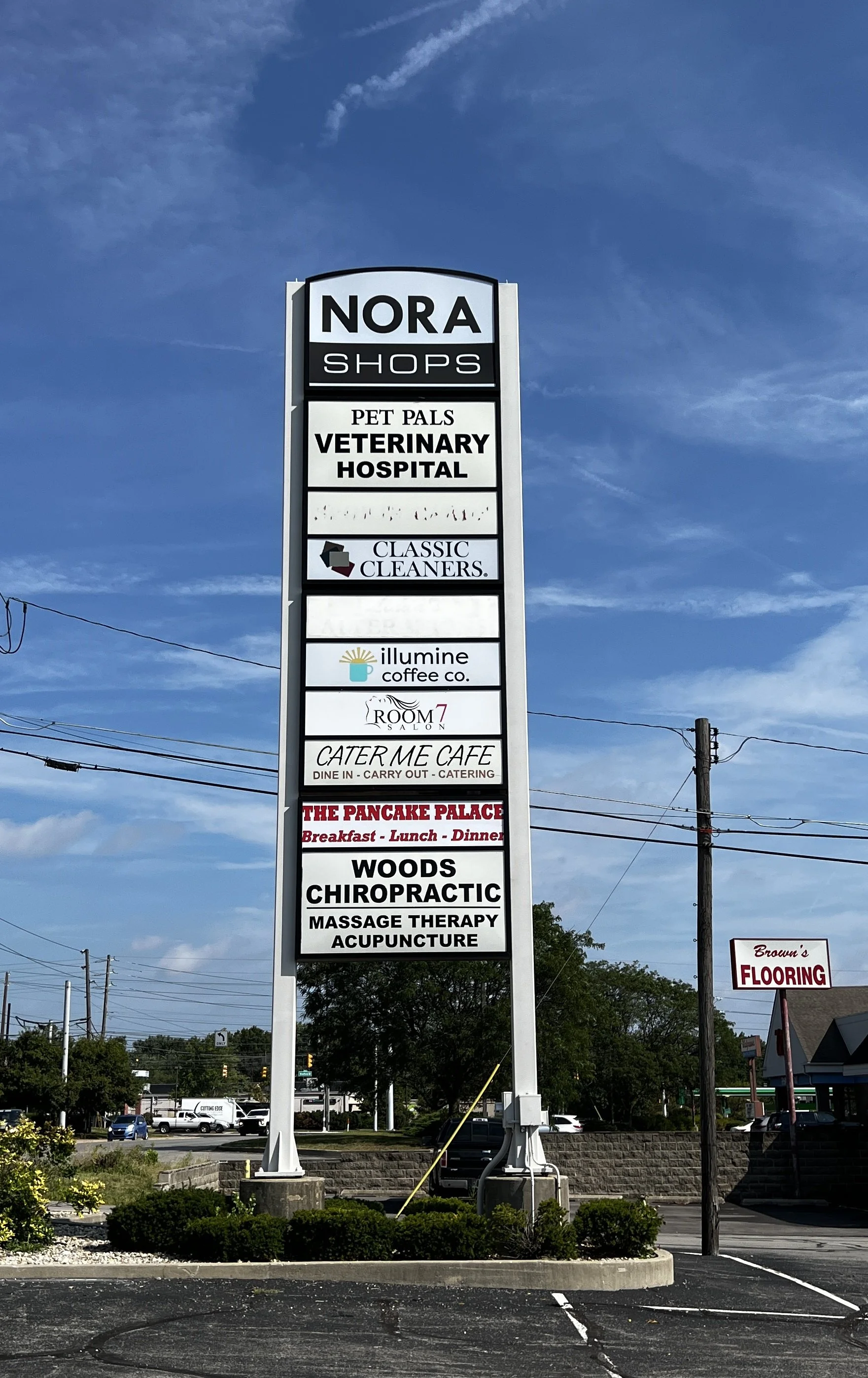 A tall vertical signboard listing various businesses, including a pet store, veterinary hospital, cleaners, coffee shop, cafe, pancake restaurant, chiropractor, massage therapy, and acupuncture, against a blue sky with some clouds and a few buildings