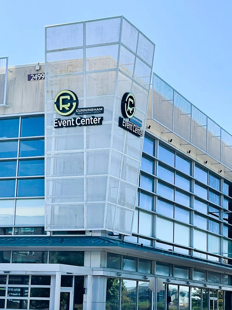 Modern multi-story building with signs for Cunningham Restaurant Group Event Center at the corner, with blue-tinted windows and clear sky