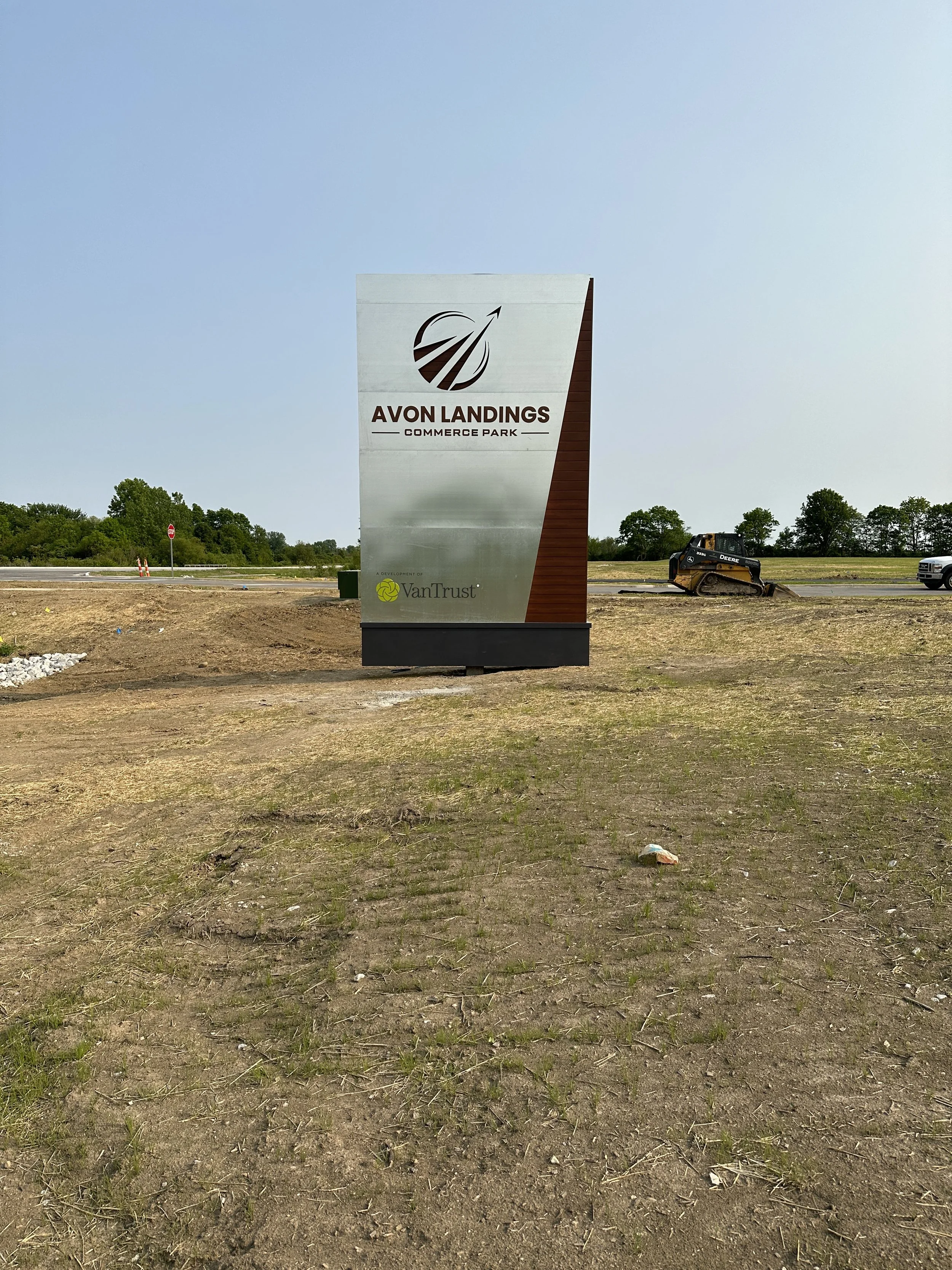 Sign for Avon Landings Commerce Park with logo and VanTrust branding, located in a grassy and dirt area with construction equipment and vehicles in background under a clear sky.