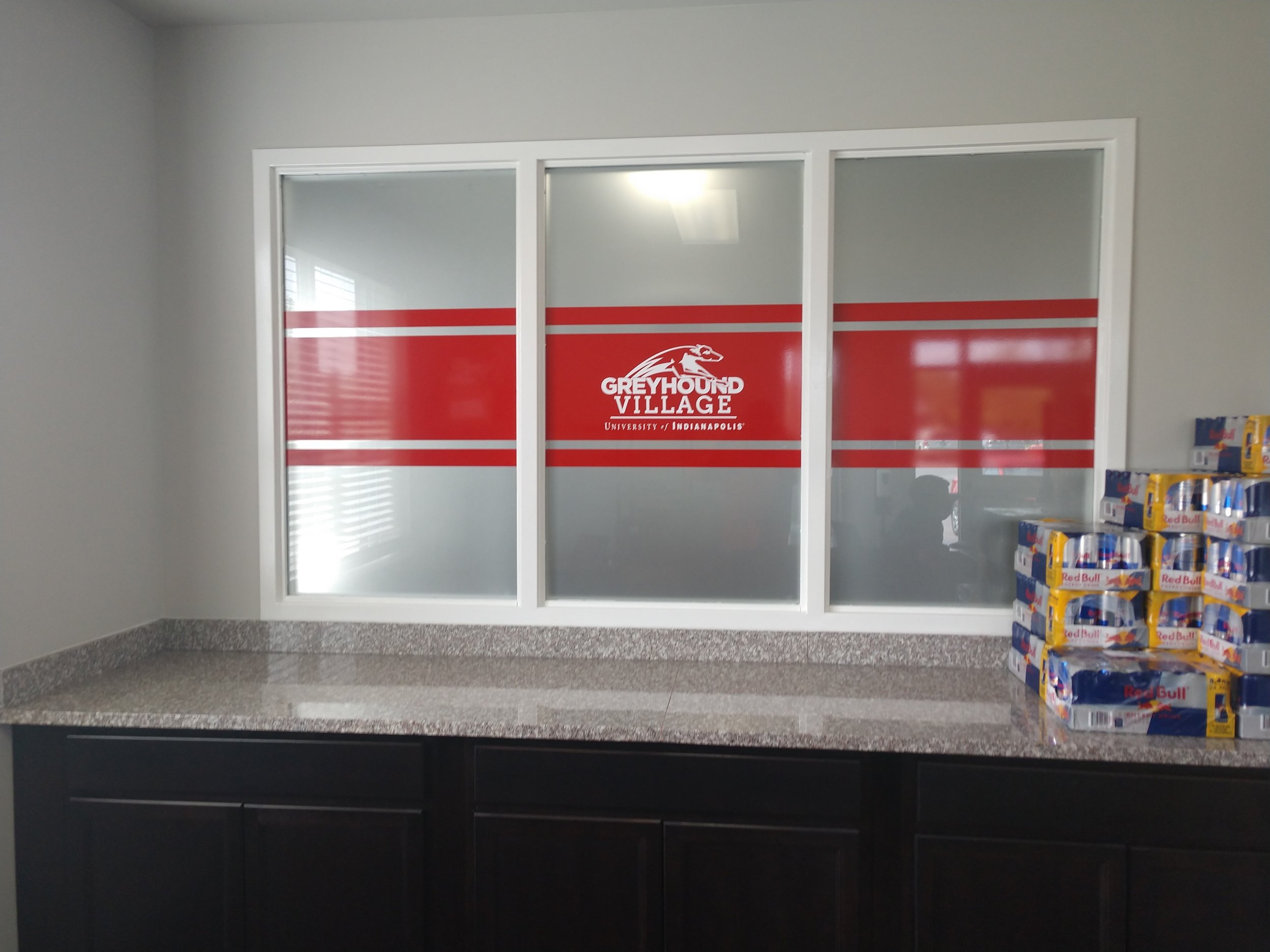 Interior of an office kitchenette with a granite countertop, a window with a red and white logo for Greyhound Village at University of Indianapolis, and a pile of Red Bull energy drink packs on the right side of the counter.