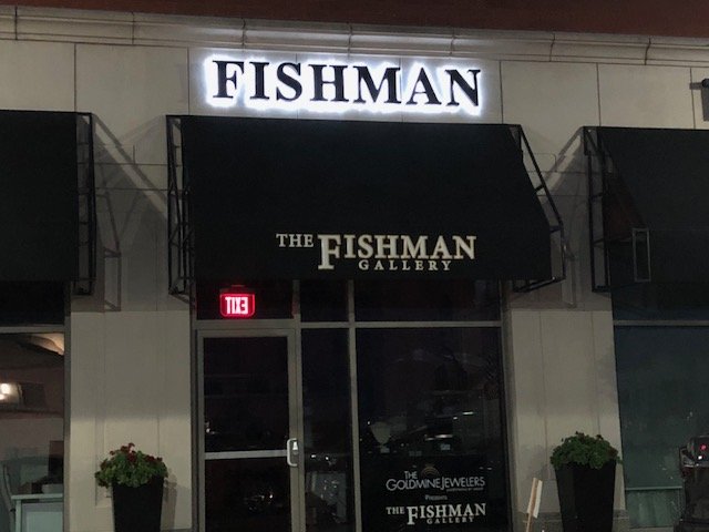 Night view of the Fishman Gallery storefront with illuminated sign and black awning. Two large potted plants on either side of the entrance.