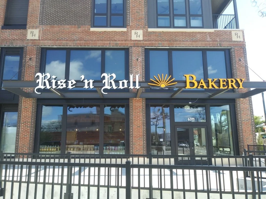 Exterior view of a bakery named 'Rise 'n Roll Bakery' in a brick building with large windows and a sign with white and yellow lettering and a sun icon.