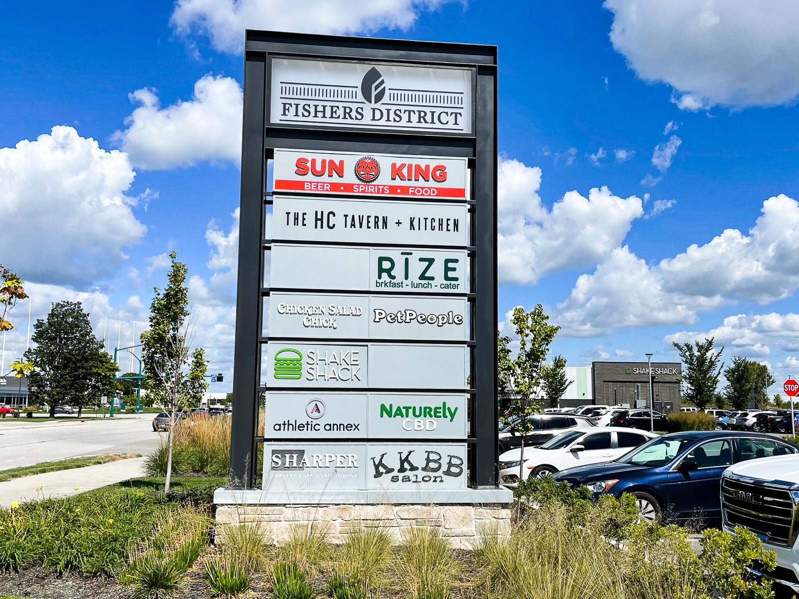 A large outdoor directory sign listing various businesses in the Fishers District shopping area, including restaurants, cafes, and wellness centers, with a parking lot and blue sky with clouds in the background.