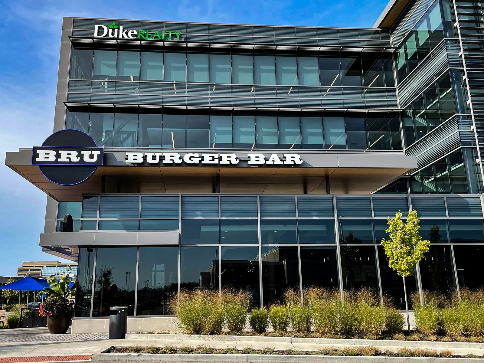 Modern multi-story building with signs for Duke Realty and Bru Burger Bar, featuring glass windows, outdoor landscaping, and a clear blue sky.