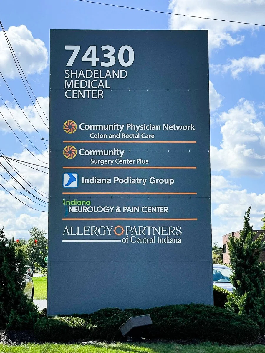Sign for 7430 Shadeland Medical Center listing various healthcare providers, including Community Physician Network, Indiana Podiatry Group, Indiana Neurology & Pain Center, and Allergy Partners of Central Indiana, with a blue sky and some trees in th