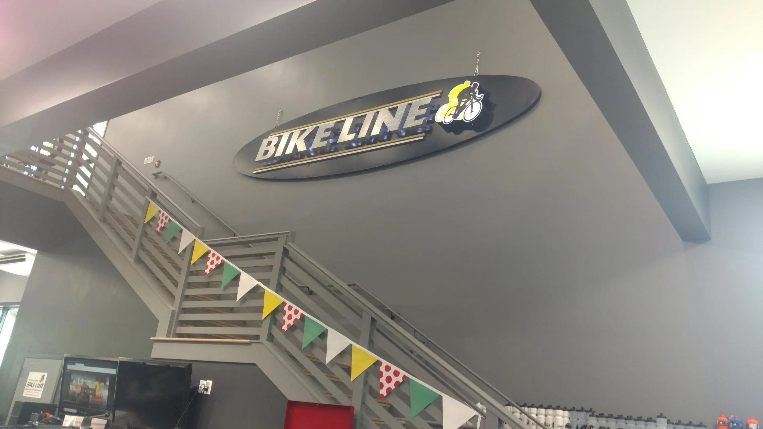Indoor view of a lobby area with a sign reading 'BIKE LINE' hanging from the ceiling, a staircase decorated with colorful pennant banners, and a counter with bottled drinks in the background.