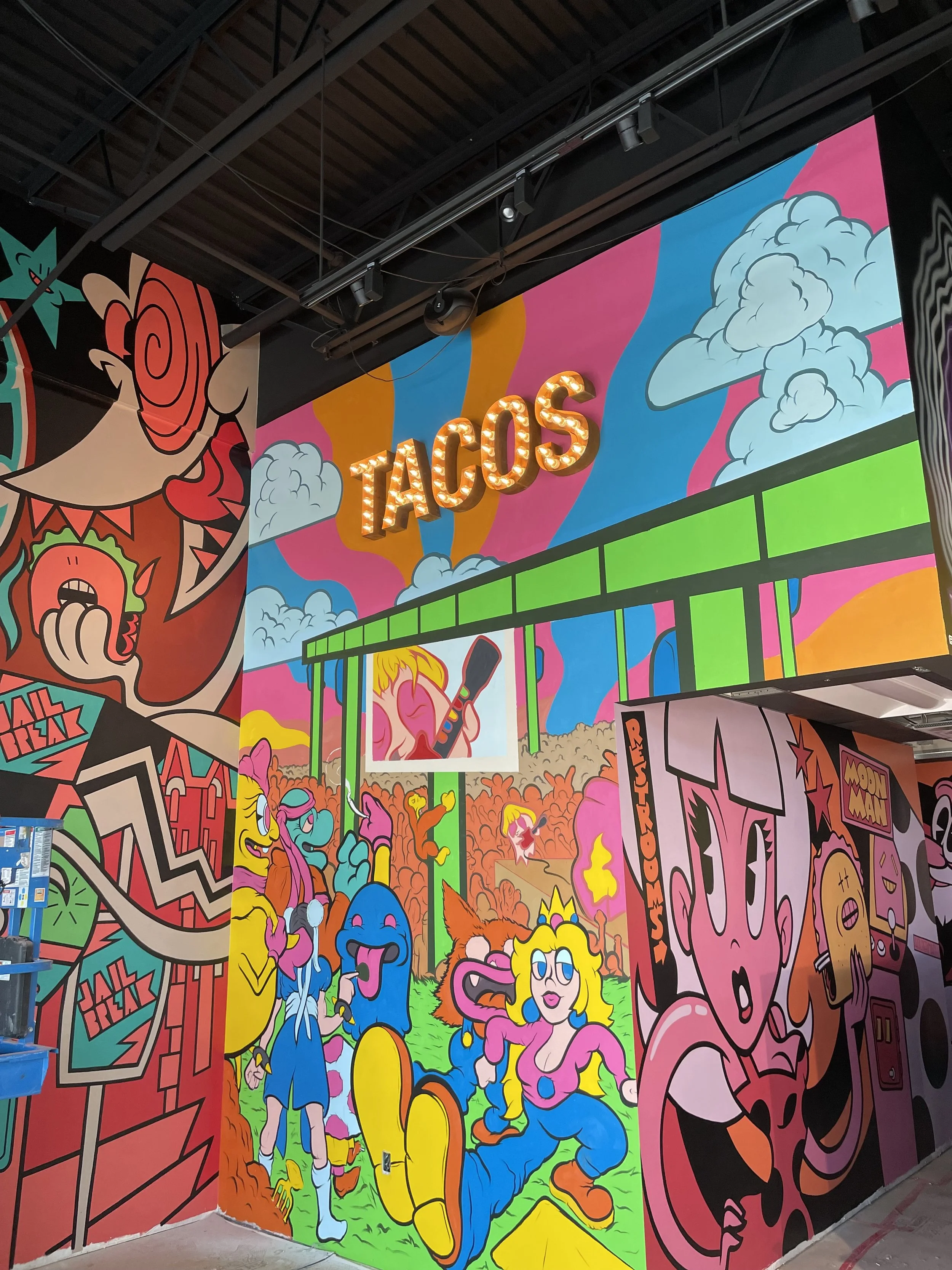 Colorful mural with various cartoon characters, clouds, and graffiti-style art, featuring a sign that says 'TACOS' with illuminated letters.