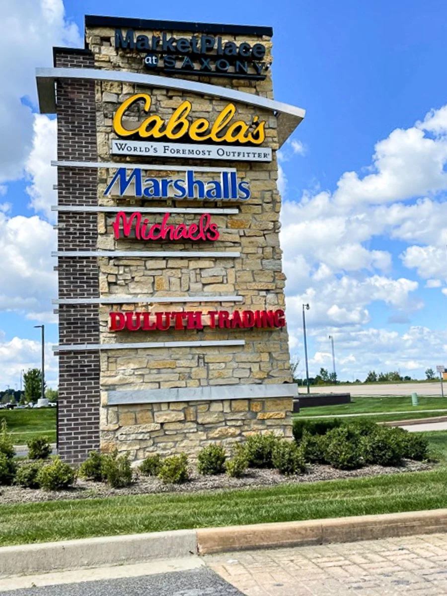 A stone sign with multiple store signs including Cabela's, Marshalls, Michaels, Duluth Trading, and MarketPlace at Saxony, situated on a grassy area with a parking lot and blue sky with clouds in the background.