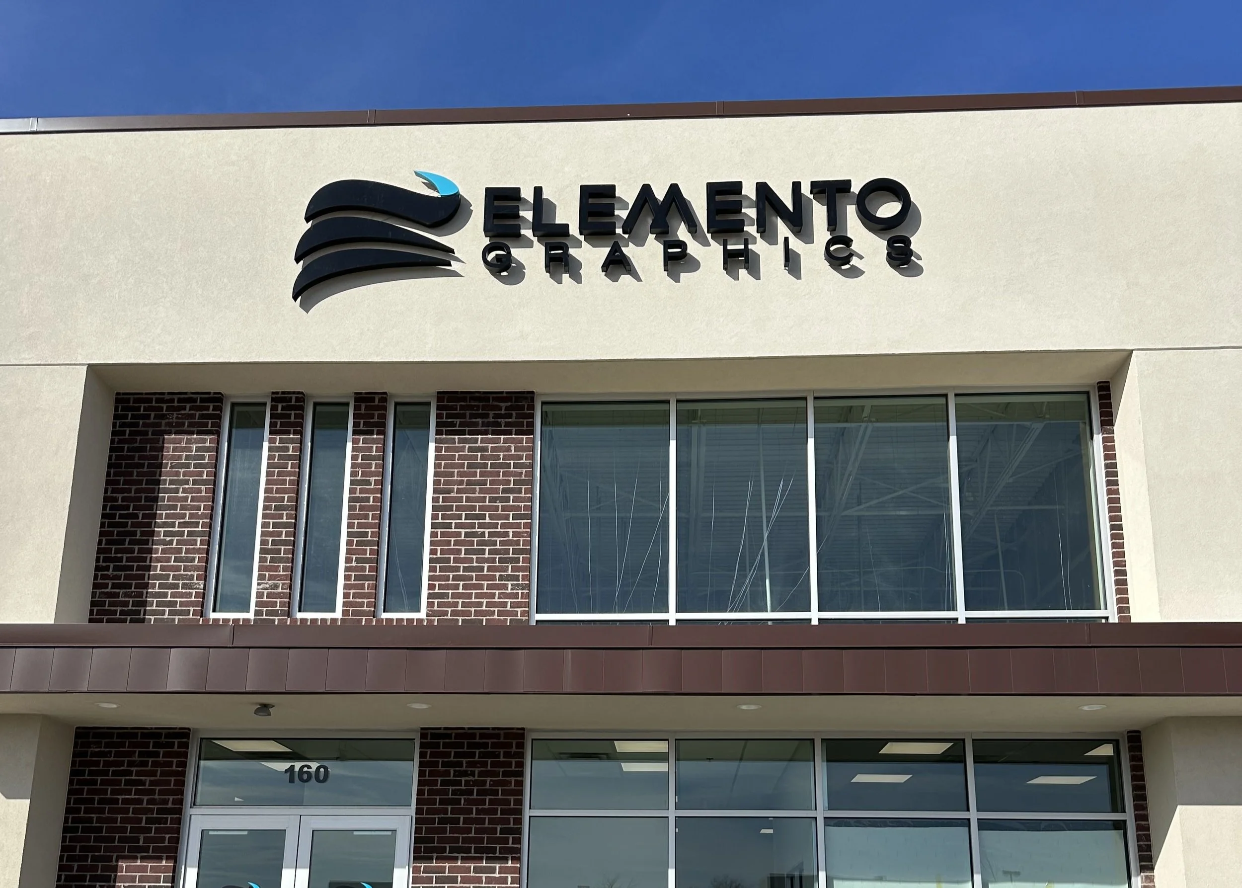 Sign on the building that reads 'ELEMENTO GRAPHICS' with a logo featuring three black curved lines and a small blue detail on the top curve.