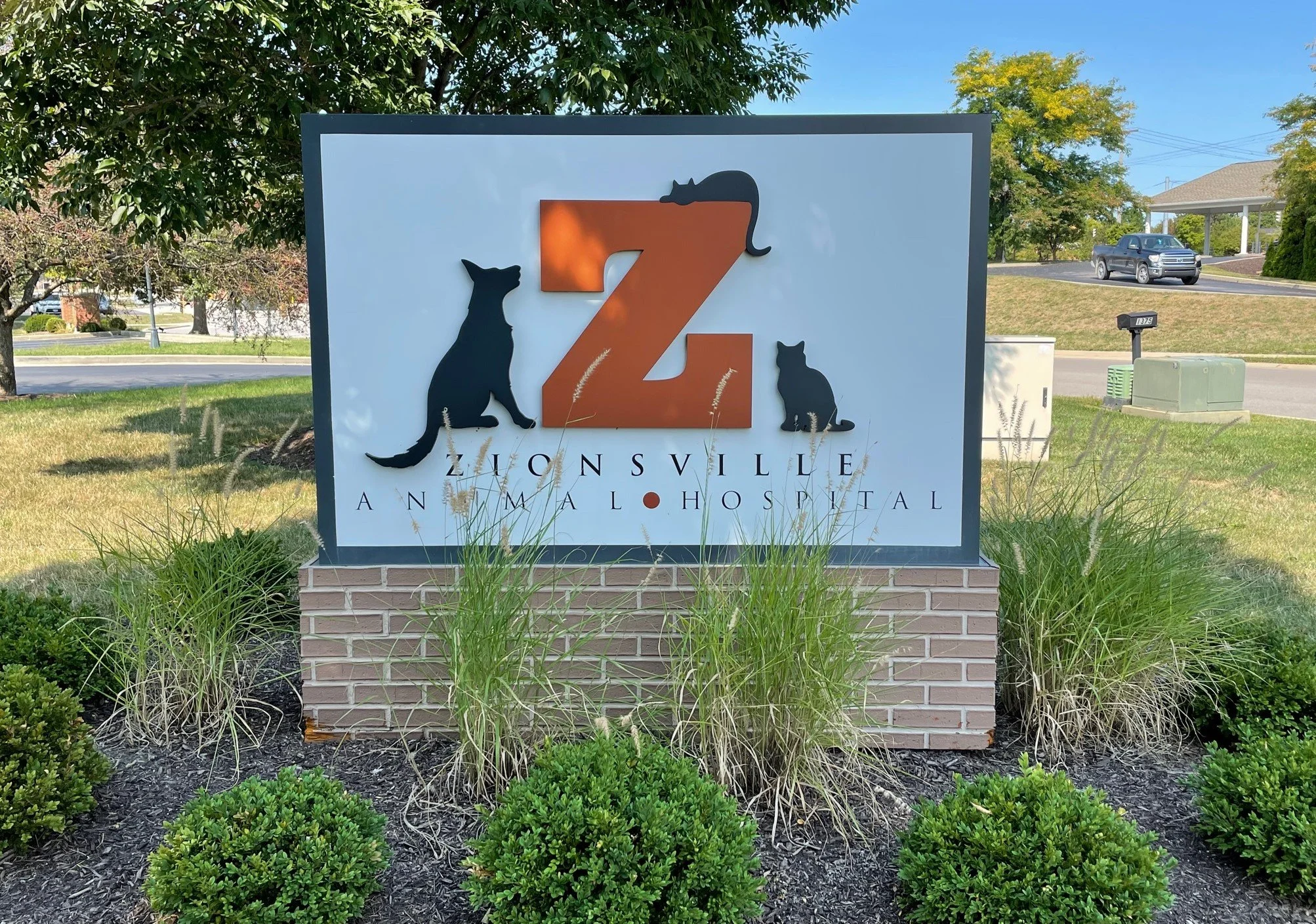 Sign for Zionsville Animal Hospital with a large orange Z and silhouettes of a cat and a dog in front of trees and a street with cars.