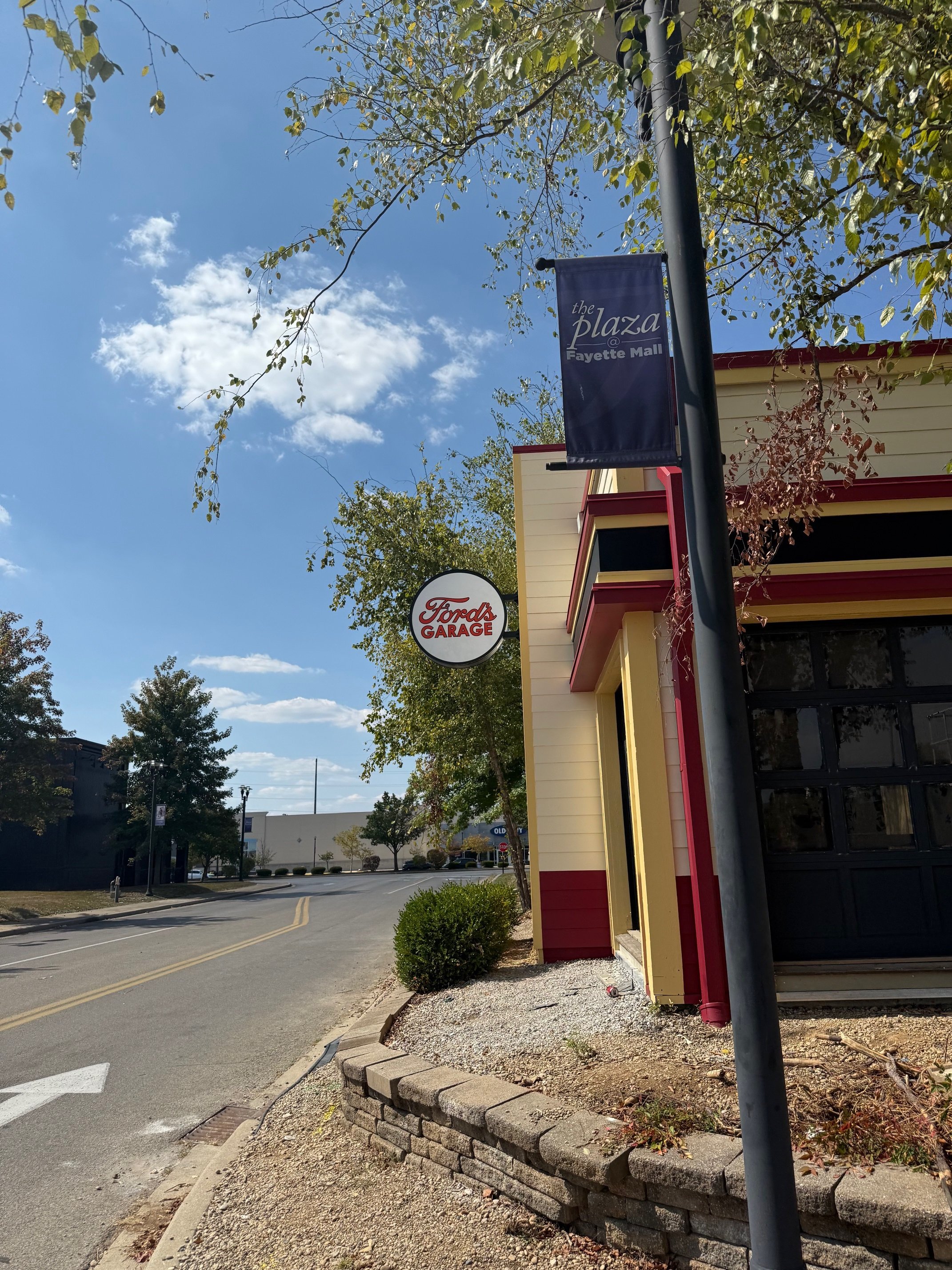 Street view with a building that has a Ford's Garage sign and a banner for the plaza at Fayette Mall, trees, and a clear blue sky.
