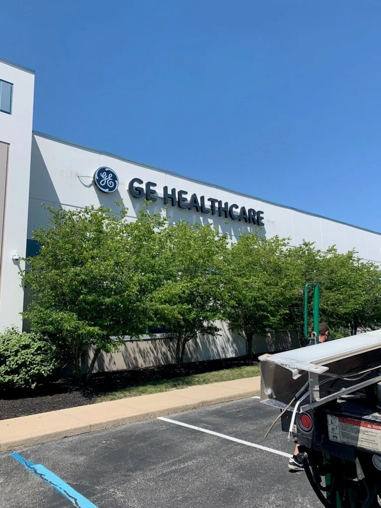 Front of a GE Healthcare building with a GE logo, trees in front, and parking lot in foreground.