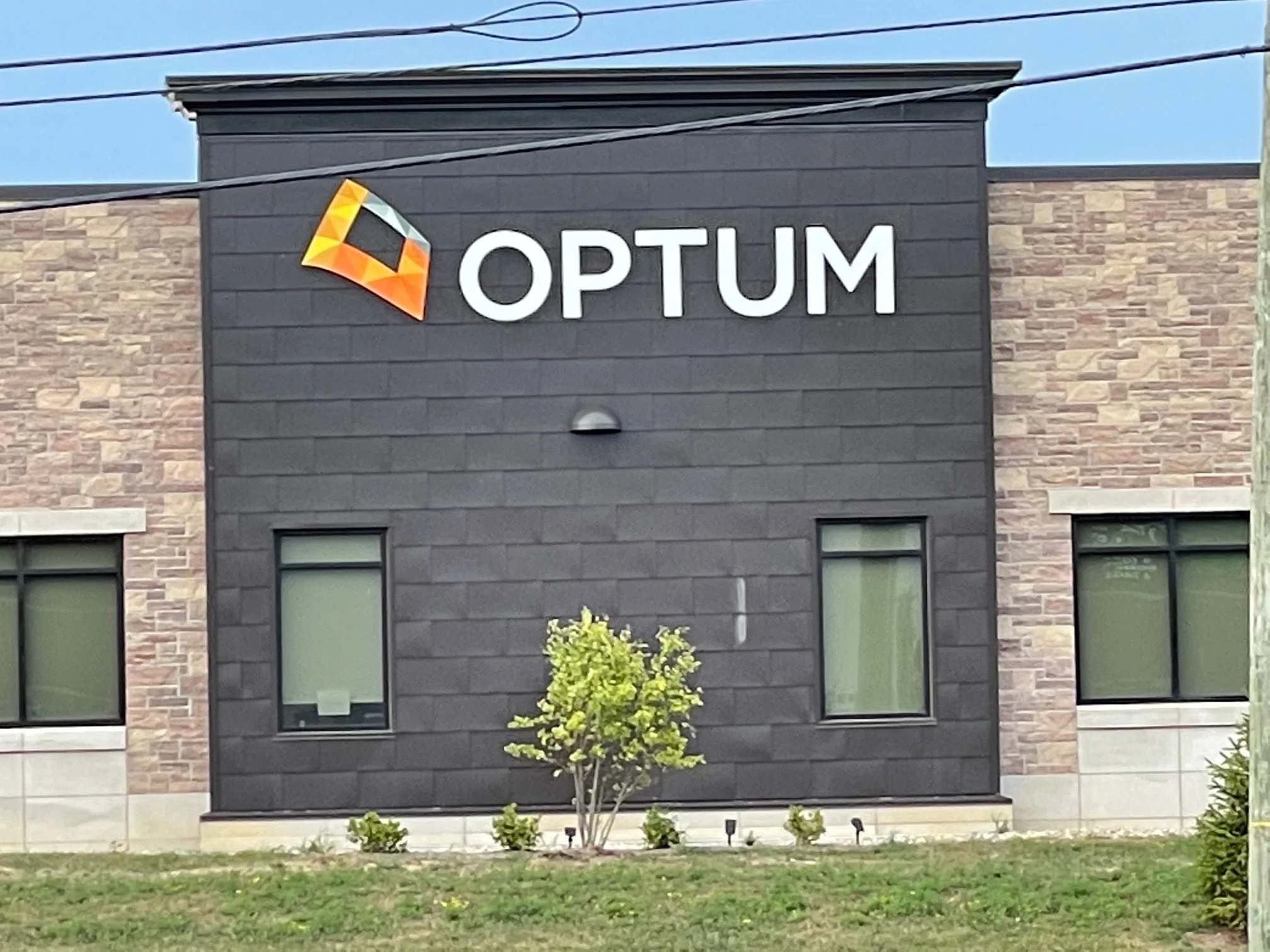 Exterior of a modern building with the sign 'OPTUM' and a colorful logo on a black facade, with windows and a small tree in front.