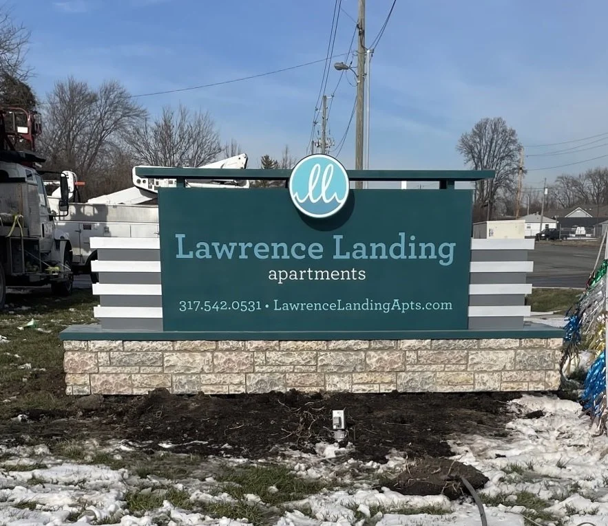 Sign for Lawrence Landing Apartments on a landscaped area with snow and patches of dirt, surrounded by trees, electrical poles, and nearby buildings.