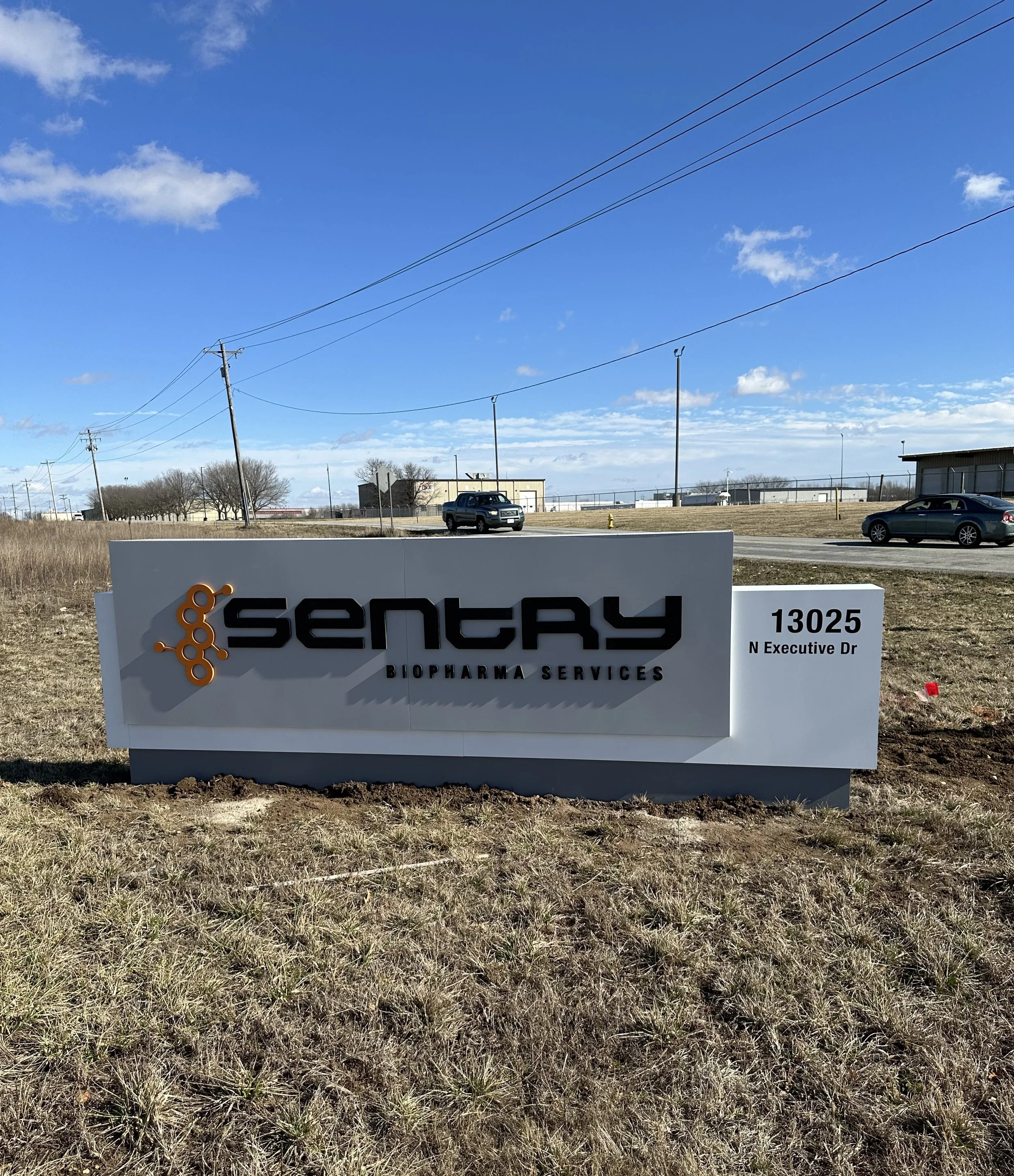Sign for Sentary Biopharma Services at 13025 N Executive Dr, with a grassy area and a parking lot in the background under a blue sky with clouds.