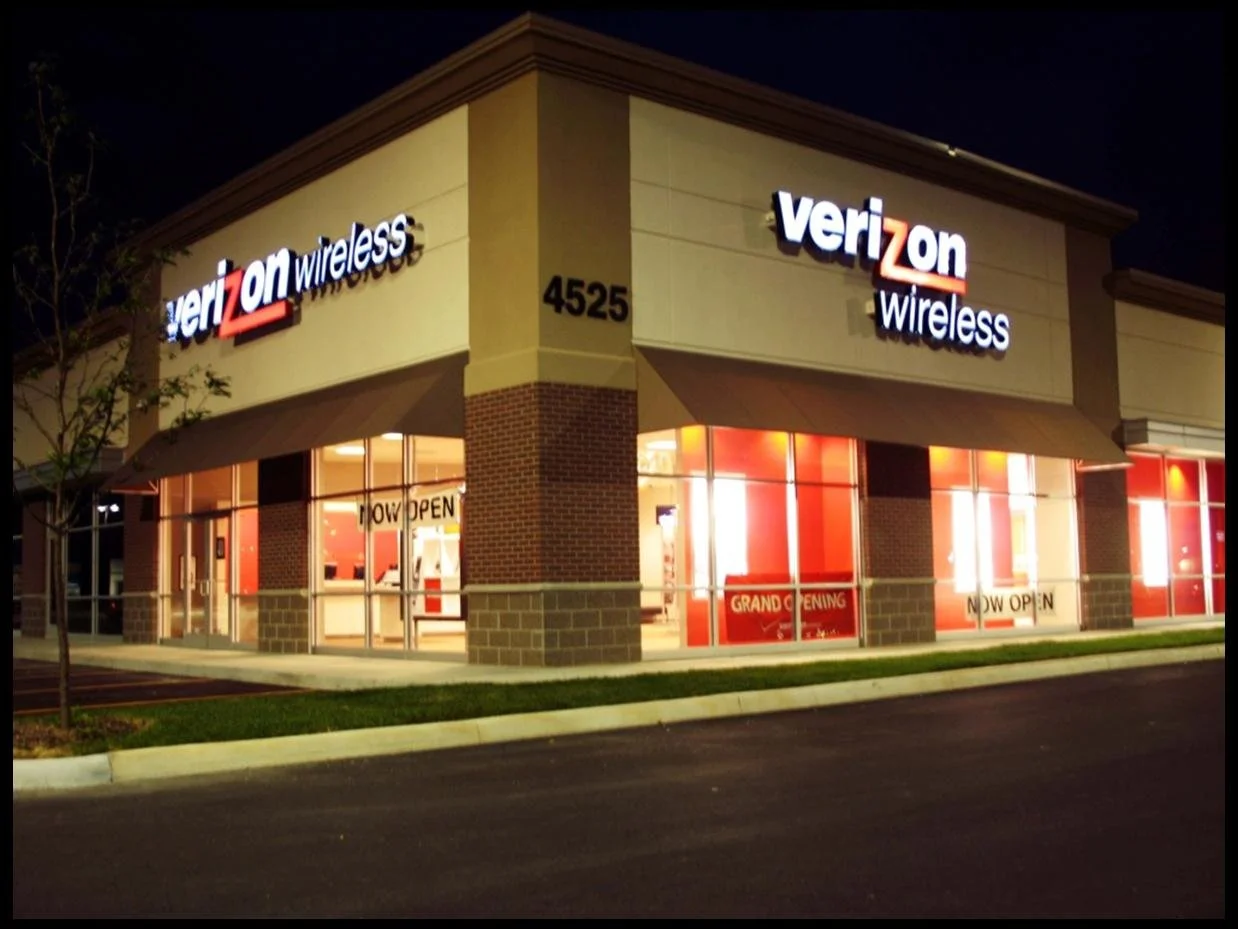 Nighttime exterior view of a Verizon Wireless store with illuminated signage, large windows displaying promotional signs, and a 'Grand Opening' banner inside.