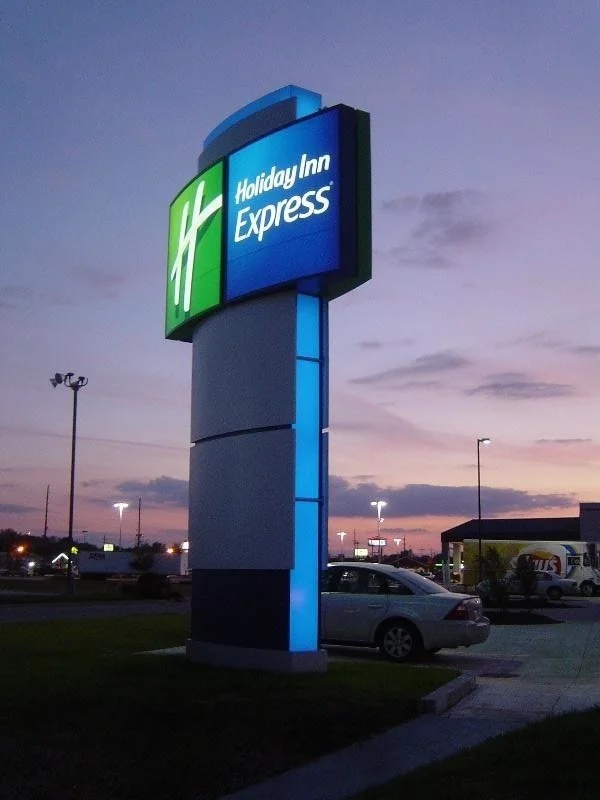 A large illuminated sign for Holiday Inn Express during dusk, with a green and blue logo and a parking lot with cars underneath.