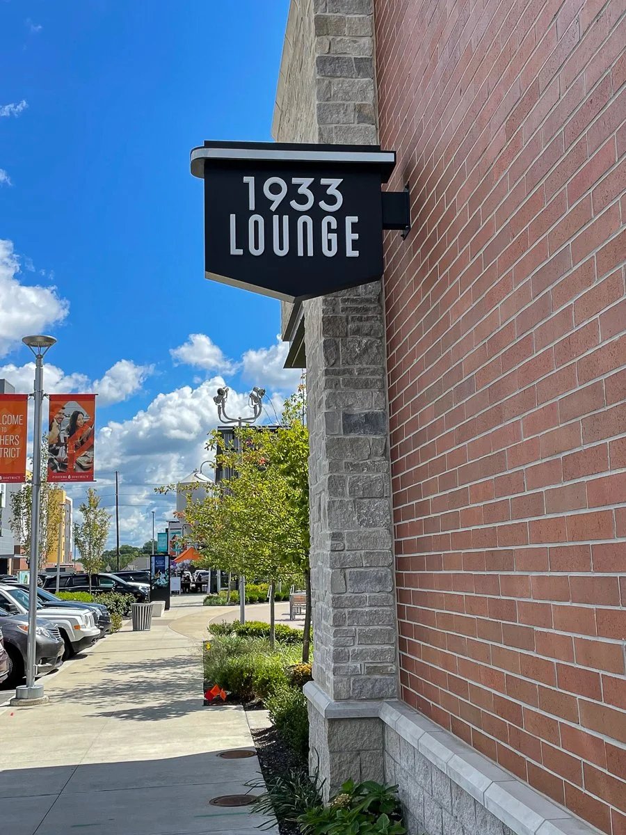 Black sign with white text reading '1933 LOUNGE' attached to a brick building exterior against a bright blue sky with a few clouds.