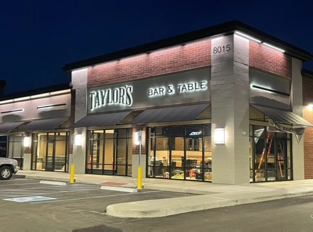Exterior of a restaurant called Taylors Bar & Table in a shopping plaza at night, with parking lot in front.