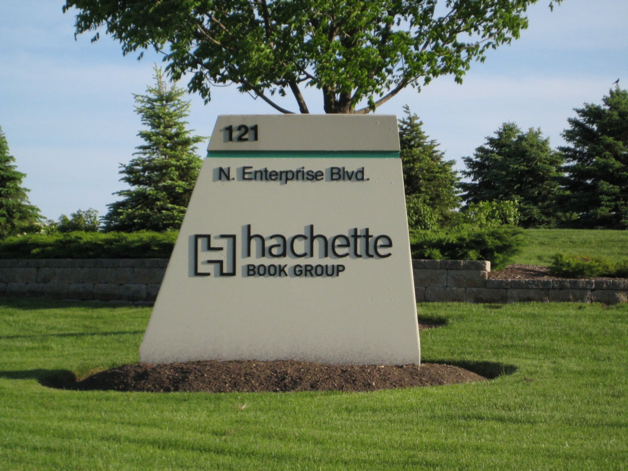 Outdoor sign for Hachette Book Group with the address 121 N. Enterprise Blvd, set in front of a landscaped area with trees and grass.