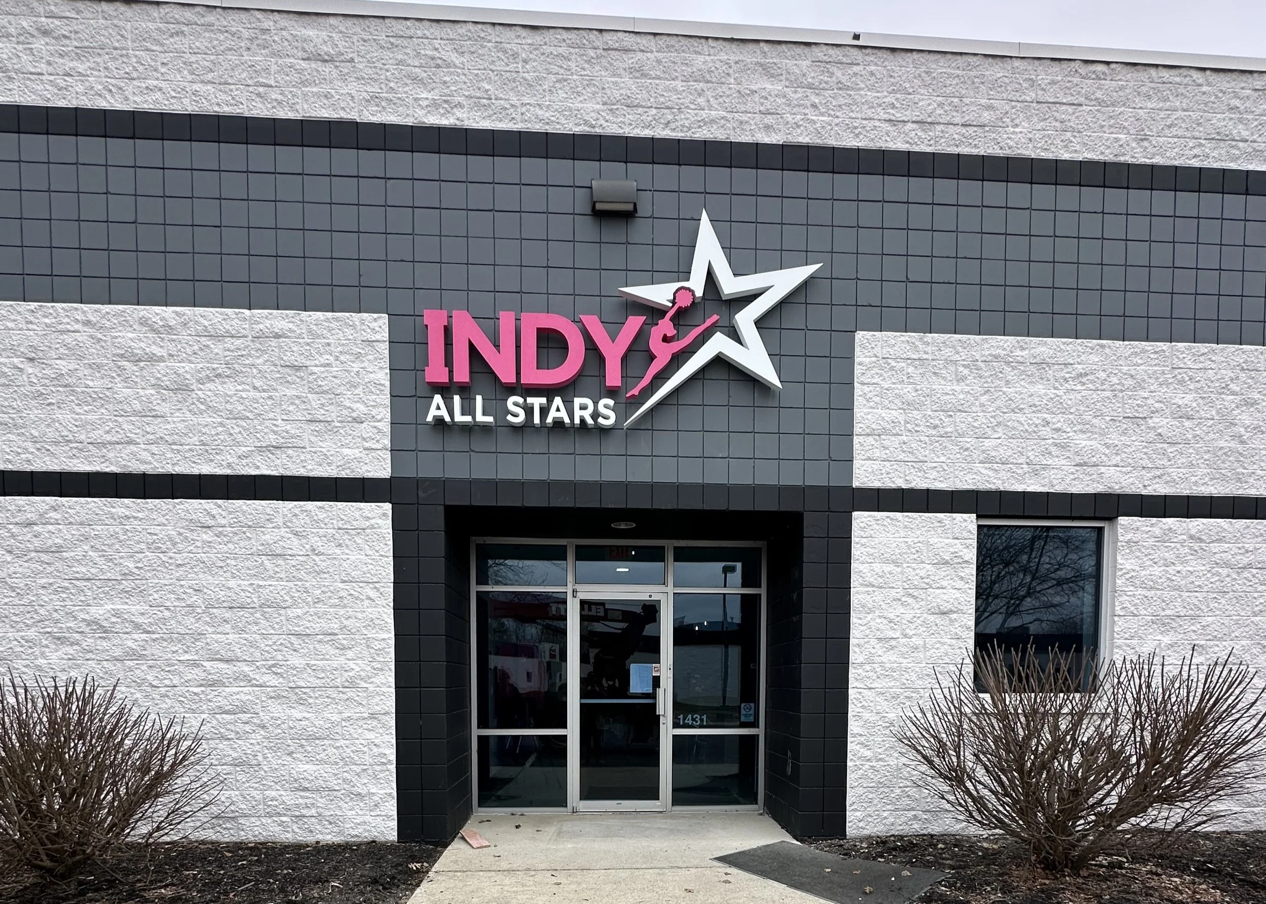 Exterior of an Indy All Stars sports facility, featuring a sign with a star and a gymnast silhouette, on a building with white and black textured walls, and a glass door entrance.