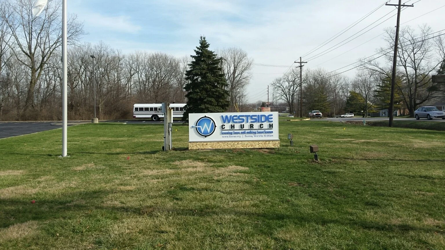 Sign in front of Westside Church with logo, grass, trees, parked bus, and cars in the background on a sunny day.