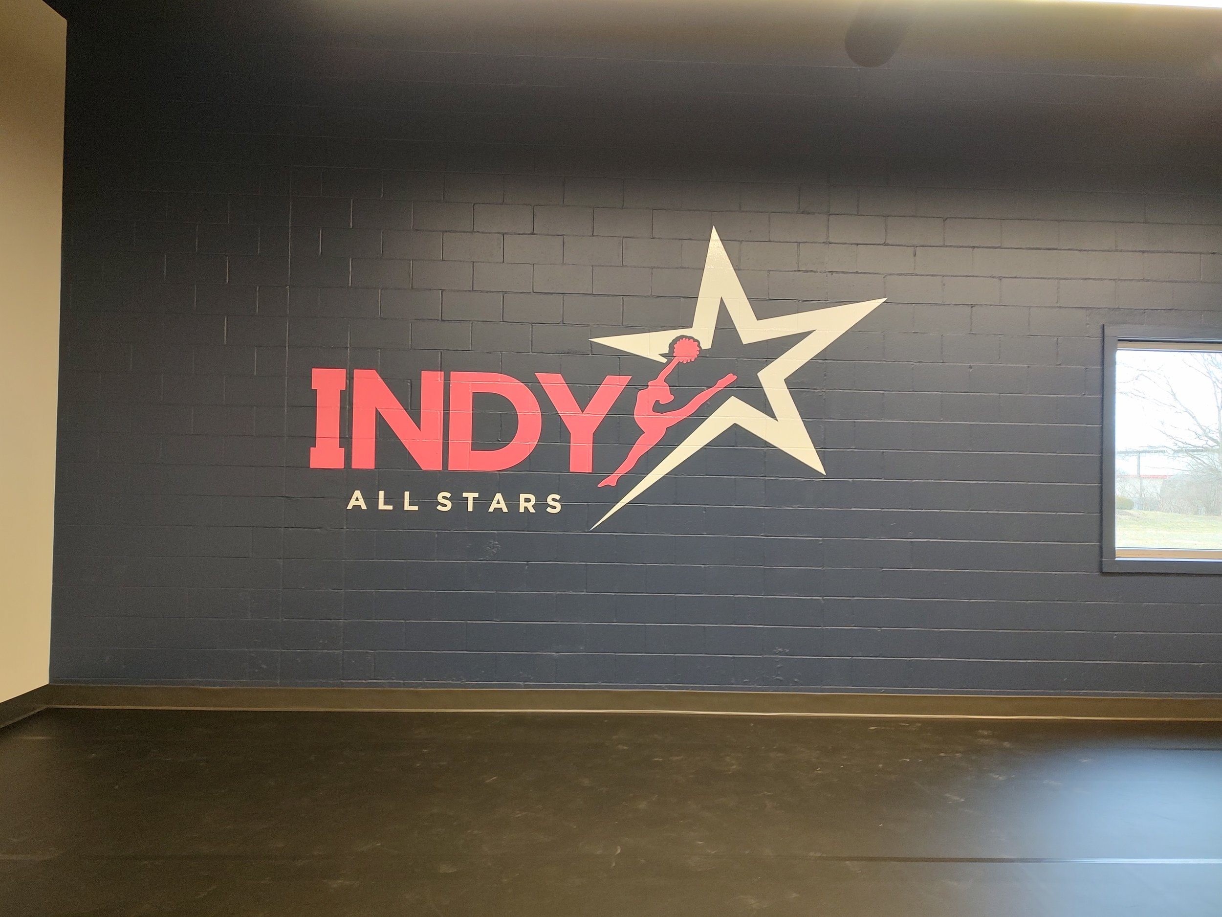 Gym wall with logo for 'Indy All Stars' featuring a star, a running figure holding a pom-pom, and bold red and white text.