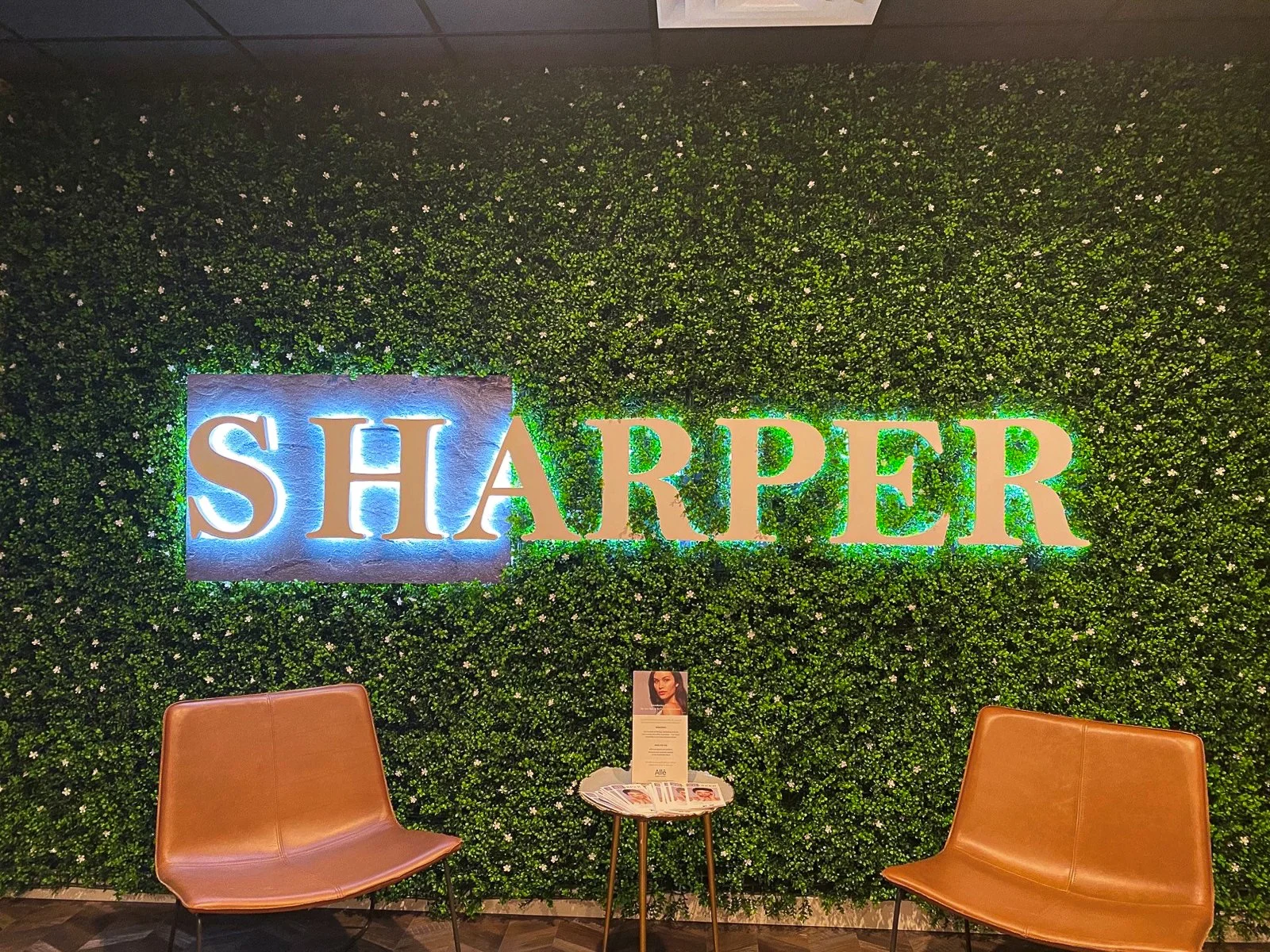 Interior of a modern space with a green wall of dense small leaves and white flowers, featuring a large illuminated sign that reads 'SHARPER'. There are two tan chairs with metal legs and a small round table between them holding informational pamphle