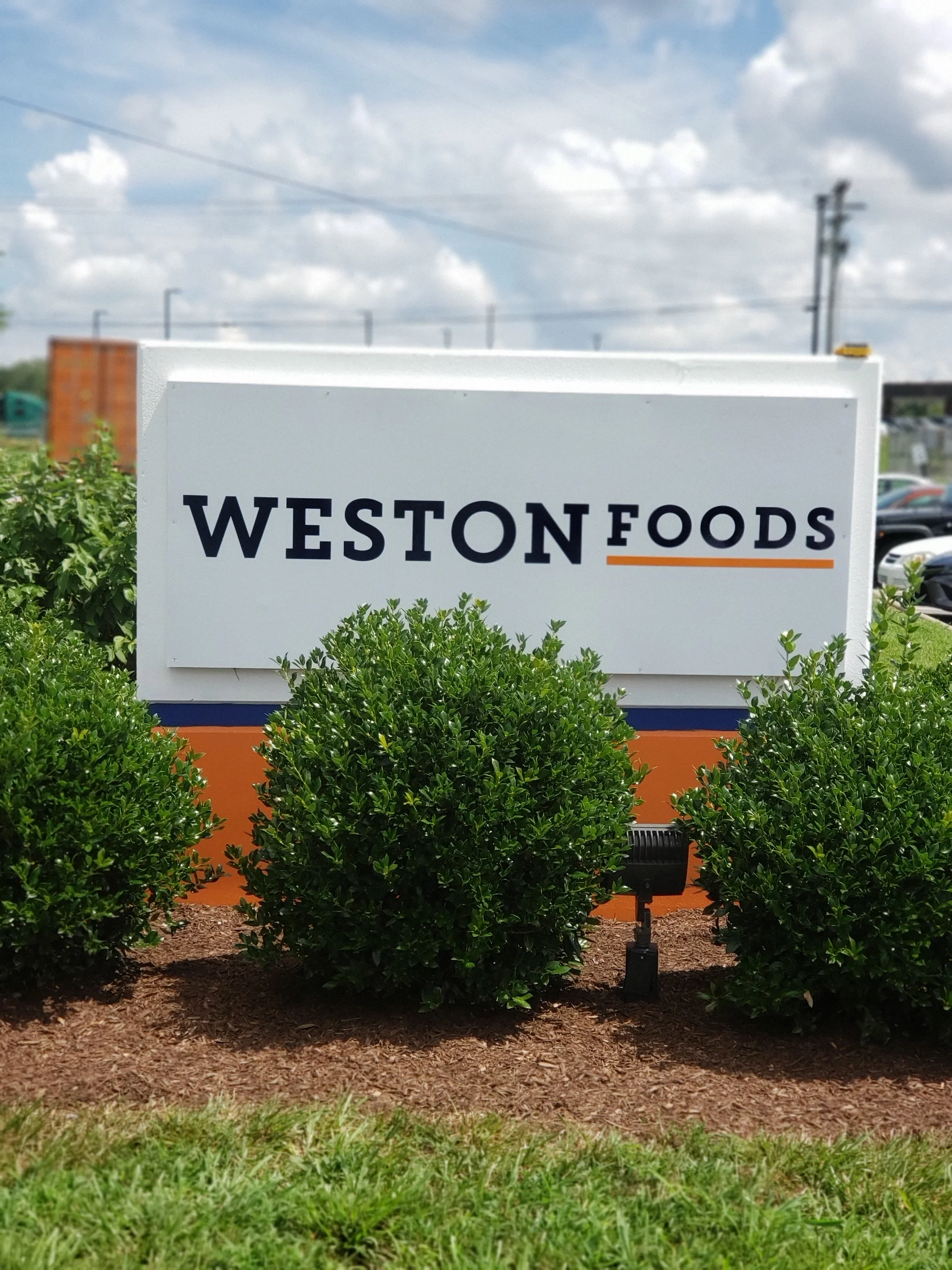 Sign for Weston Foods with bushes in front and a partly cloudy sky