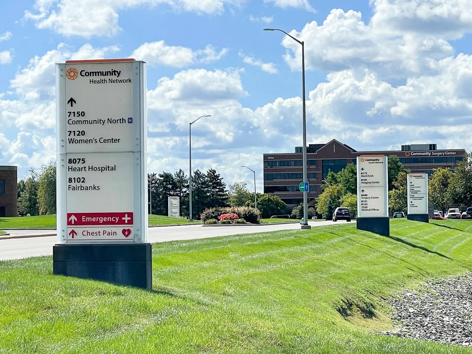A signboard for Community Health Network with directions to various medical facilities including Community North, Women's Center, Heart Hospital, and Fairbanks. In the background, there are additional signs, parking lot, trees, and a large multi-stor