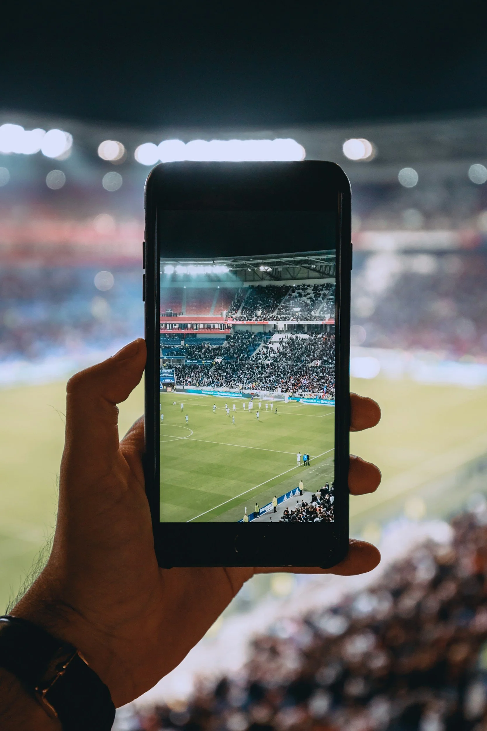 A person holding a smartphone taking a photo of a soccer game in a packed stadium at night.