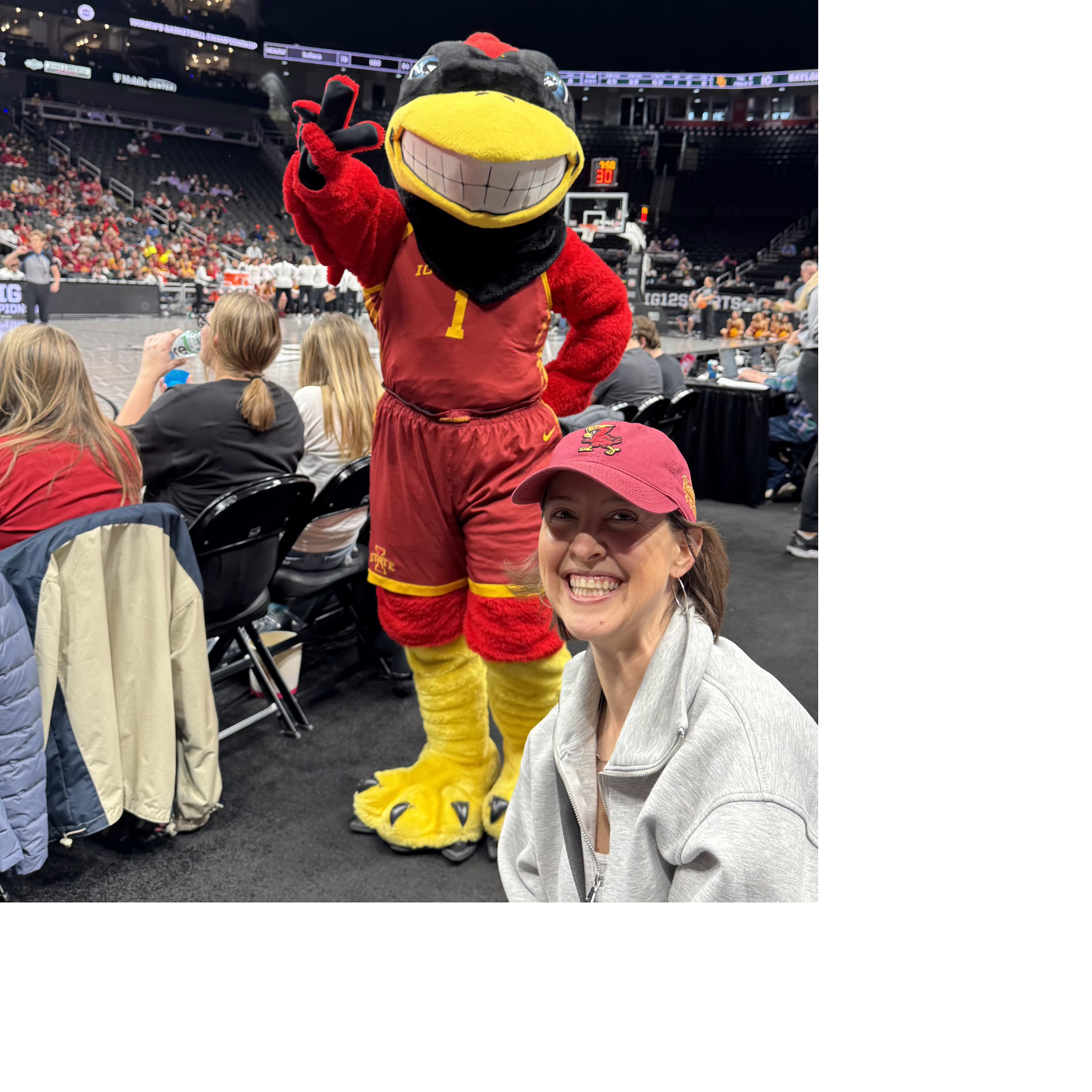 Jenny smiling and sitting courtside at a basketball game with Cy the Cyclone mascot standing behind her. The mascot is waving and making a peace sign. The background shows spectators and the indoor basketball court.