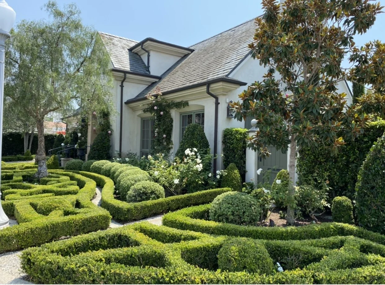 Well-maintained front yard garden with hedges, bushes, and trees in front of a white house with gray roof.