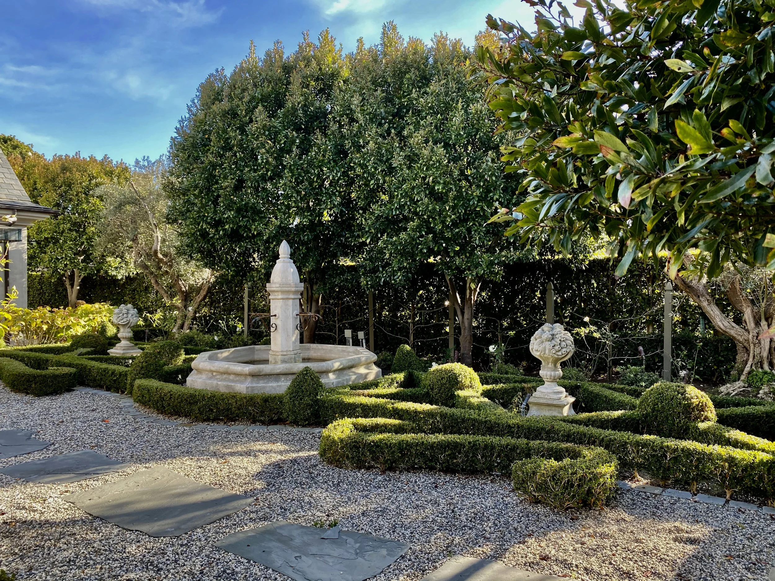 A landscaped backyard garden featuring a stone fountain, trimmed hedges, decorative urns, and tall trees with a blue sky in the background.