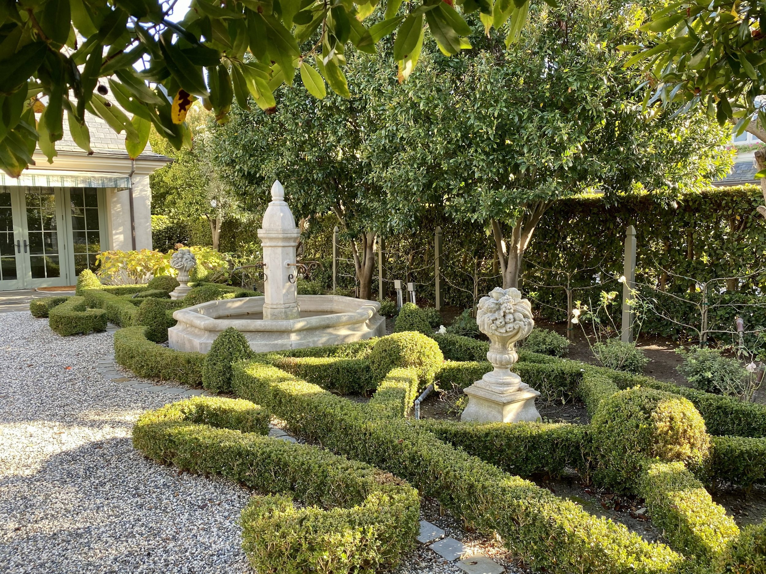 A backyard garden with a stone fountain in the center, surrounded by neatly trimmed bushes and decorative stone planters, with trees and a house with large glass doors in the background.